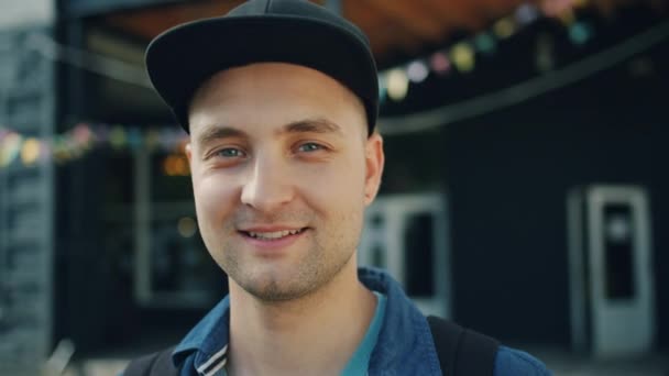 Lähikuva muotokuva hymyilevä nuori kaveri yllään trendikäs lippis seisoo ulkona — kuvapankkivideo