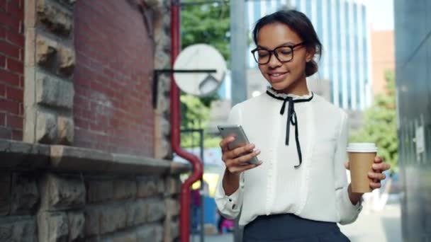 Leende afrikansk amerikansk kvinna med hjälp av smartphone hålla kaffe gå utomhus — Stockvideo