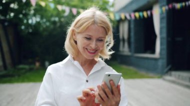 Açık havada gülümseyen ekrana bakan akıllı telefon kullanan neşeli kadın