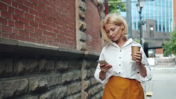 Улыбающаяся взрослая женщина использует смартфон холдинг взять кофе ходить на улицу — стоковое видео