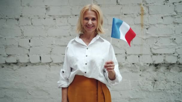 Atractiva mujer madura ondeando bandera francesa al aire libre sobre fondo de pared de ladrillo — Vídeos de Stock