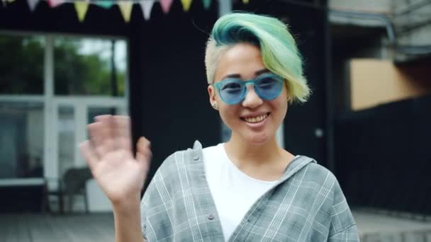 Азиатская юная леди с окрашенными волосами машет рукой, улыбаясь стоя на улице — стоковое видео