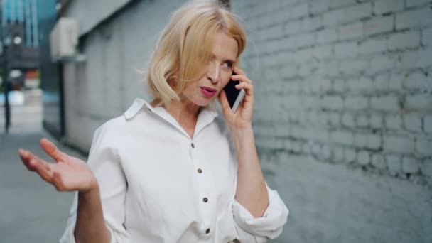 Slow Motion van vrolijke rijpe vrouw praten op mobiele telefoon staande buitenshuis — Stockvideo