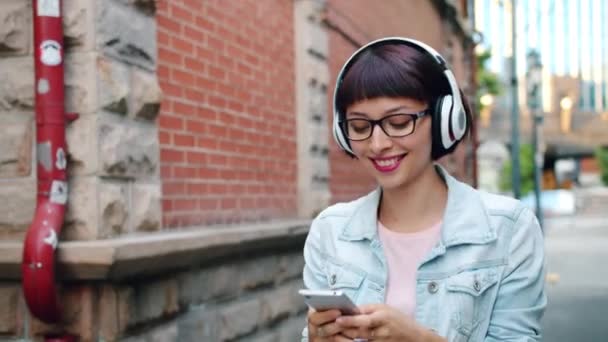 Красива леді слухає музику в навушниках, використовуючи смартфон, що йде по вулиці — стокове відео