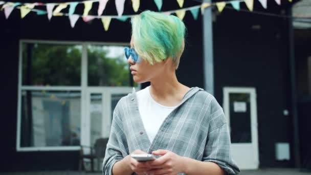 Retrato de una chica punk bastante asiática usando la pantalla táctil del teléfono inteligente al aire libre — Vídeos de Stock