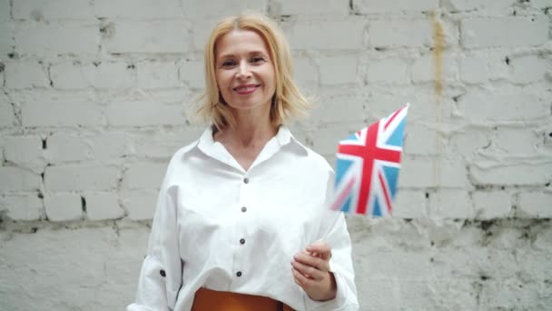 Hermosa señora de mediana edad ondeando bandera británica sonriendo en el fondo de la pared de ladrillo — Vídeo de stock