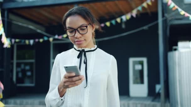 Hermosa mujer de raza mixta usando teléfono inteligente sonriendo pantalla táctil al aire libre — Vídeo de stock