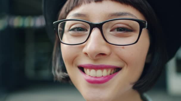 Retrato de cerca de una bonita adolescente con gafas y sombrero sonriendo al aire libre — Vídeos de Stock