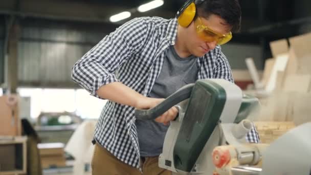 Ξυλουργός σε γυαλιά και ακουστικά που εργάζονται με ηλεκτρικό πριόνι στο εργαστήριο — Αρχείο Βίντεο