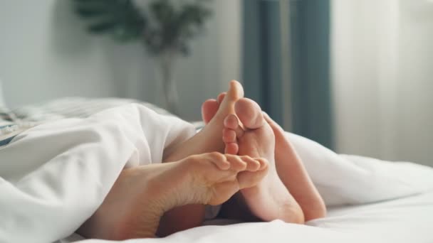 Pies desnudos humanos tocándose en la cama bajo una manta blanca en el dormitorio — Vídeos de Stock