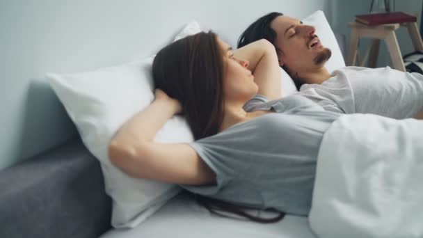 Раздраженная женщина покрывает голову подушкой, когда парень храпит во сне в постели — стоковое видео