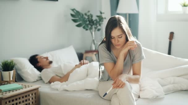 Нервова дівчина сидить на ліжку тримає тест на вагітність, коли хлопець спить у спальні — стокове відео