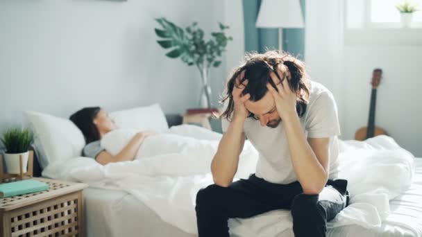 Unglücklicher Ehemann sitzt auf Bett und berührt trauriges Gesicht, während Frau zu Hause schläft — Stockvideo