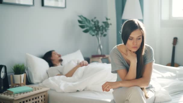 Сумна дівчина в піжамі сидить на ліжку думаючи, що зітхає, коли хлопець спить вдома — стокове відео