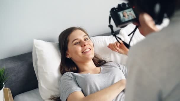 Aantrekkelijk meisje poseren voor camera liggend in bed, man met professionele camera — Stockvideo