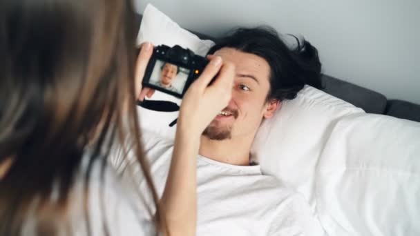 Juguetón joven posando en casa en la cama mientras niña utilizando la cámara moderna — Vídeos de Stock