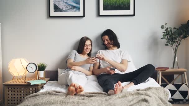 Make och fru använder smartphone skrattar sitter i sängen hemma — Stockvideo