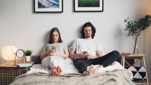 Chica y chico usando teléfonos inteligentes sentados en la cama en casa con caras serias — Vídeos de Stock