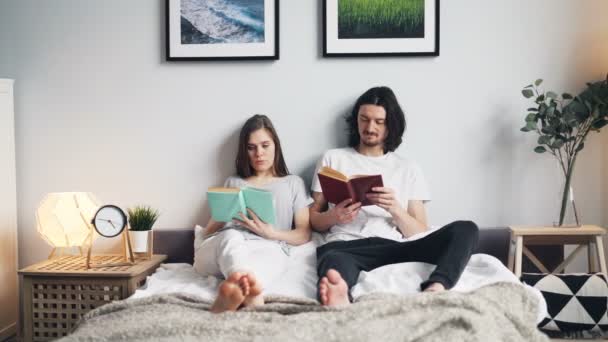 Портрет девушки и парня, читающих книги в постели дома в светлой квартире — стоковое видео