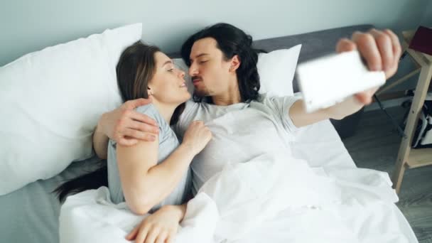 Мужчина и женщина делают селфи в постели дома, используя камеру смартфона, развлекаясь — стоковое видео