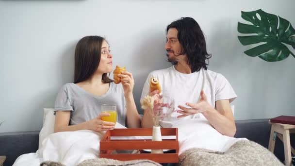 Radosna para eaing croissanty w łóżku rozmawiając i śmiejąc się rano — Wideo stockowe