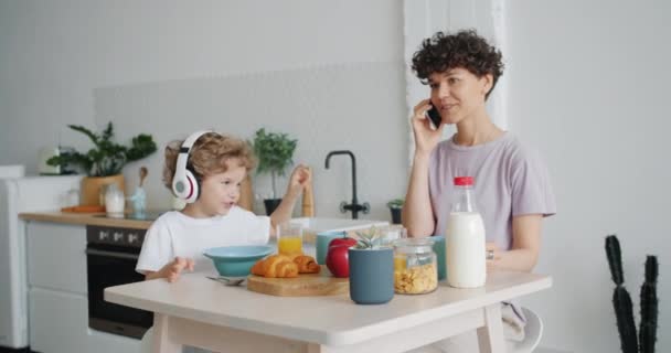 Γυναίκα μιλάει στο κινητό τηλέφωνο, ενώ ο γιος απολαμβάνει μουσική στα ακουστικά στην κουζίνα — Αρχείο Βίντεο