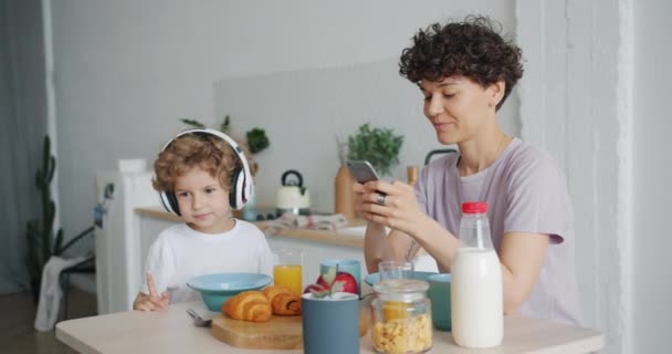 Νεαρή κυρία χρησιμοποιώντας smartphone, ενώ μικρό αγόρι απολαμβάνει μουσική στο τραπέζι της κουζίνας — Αρχείο Βίντεο