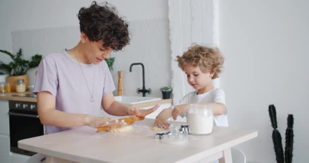 Chica feliz e hijo pequeño rodando masa haciendo galletas en casa en la cocina — Vídeos de Stock