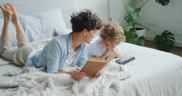 Familia feliz mamá y el niño leyendo libro y el uso de la tableta en la cama en casa — Vídeos de Stock