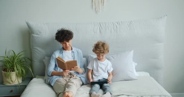 Νεαρή κυρία διαβάζοντας το βιβλίο, ενώ μικρό γιο απολαμβάνοντας tablet κάθεται στο κρεβάτι στο σπίτι — Αρχείο Βίντεο