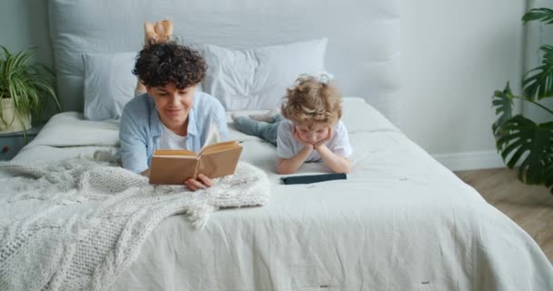 Amante mãe leitura livro, em seguida, assistindo desenhos animados com o filho usando tablet no quarto — Vídeo de Stock