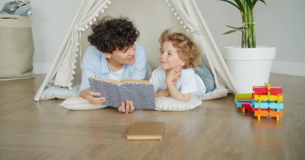 Szerető anya olvasatban könyvet aranyos gyermek feküdt a padlón hangulatos sátor bent — Stock videók