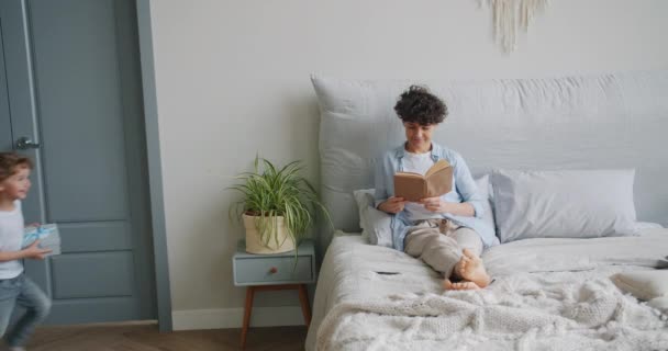 Mujer joven leyendo libro en la cama cuando el niño le da su regalo en caja de regalo — Vídeos de Stock