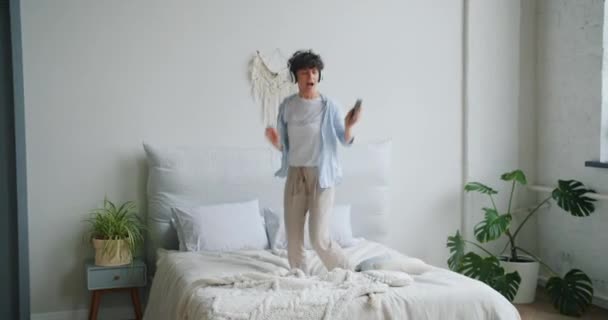 Schattig actief meisje springen dansen op bed dragen hoofdtelefoon houden smartphone — Stockvideo