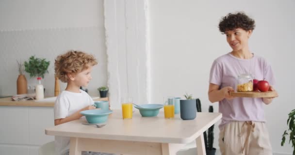 Madre sonriente trayendo cereales para el desayuno y manzanas para un niño feliz en casa — Vídeos de Stock