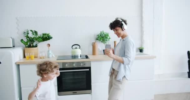 Madre en auriculares e hijo pequeño bailando en la cocina divirtiéndose juntos — Vídeo de stock