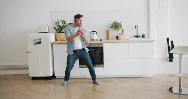 Мужчина в наушниках танцует на кухне, держа смартфон, развлекаясь дома. — стоковое видео