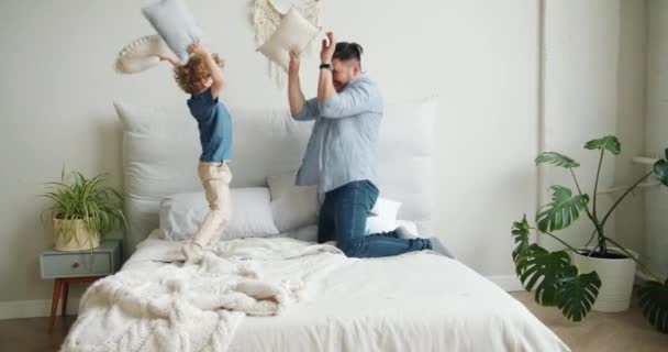 Junger Mann kämpft mit Kissen und kleinem Sohn, der Spaß auf dem Bett in der Wohnung hat — Stockvideo