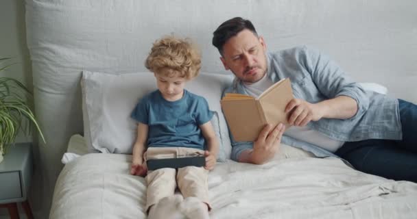 진지한 남자 독서 책 에 침대 동안 작은 아이 보고 만화 에 태블릿 — 비디오