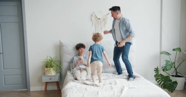 Πατέρας και γιός άλμα στο κρεβάτι στο σπίτι, ενώ η μητέρα χρησιμοποιώντας smartphone χαμογελά — Αρχείο Βίντεο