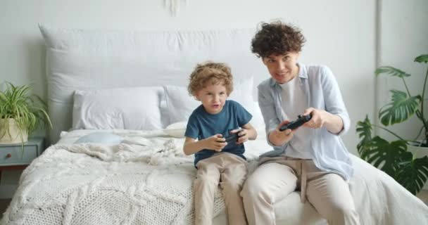 Mãe e filho jogando videogame sentado na cama no quarto em casa se divertindo — Vídeo de Stock