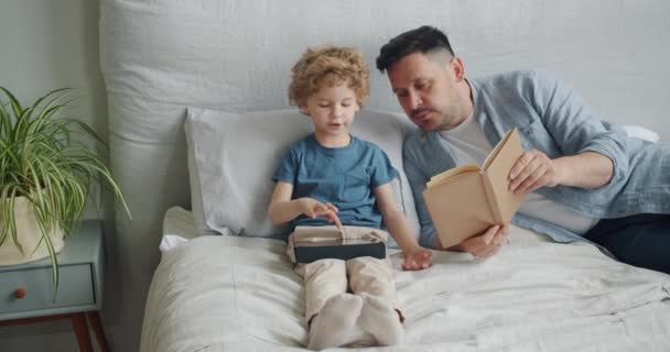 Padre amoroso leyendo libro a niño mientras niño usando tableta en la cama en el apartamento — Vídeo de stock