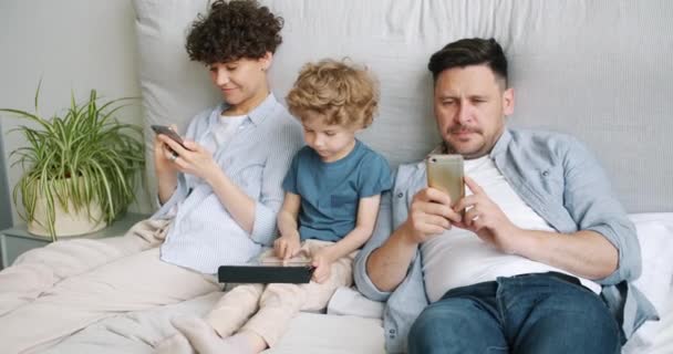 Мужчина и женщина используют смартфоны в то время как ребенок играет в игру на планшете в постели дома — стоковое видео