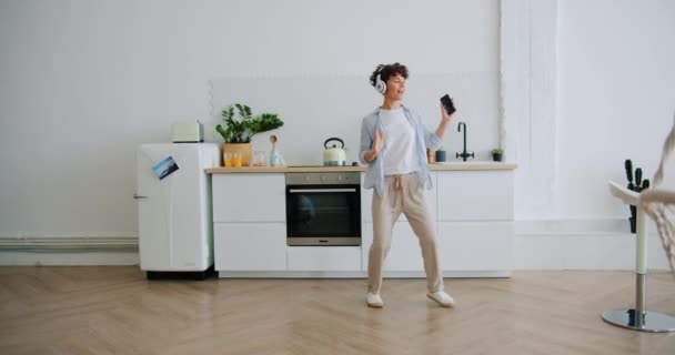 Женщина в наушниках танцует, слушая музыку, держащую смартфон на кухне — стоковое видео
