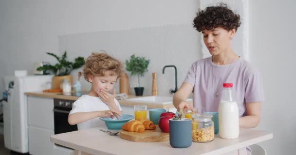 年轻女子喝橙汁，而儿子在厨房的餐桌上吃麦片 — 图库视频影像