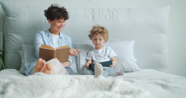 Anya olvasatban könyvet, miközben gyermek használ tablettát ágyon modern apartman — Stock videók