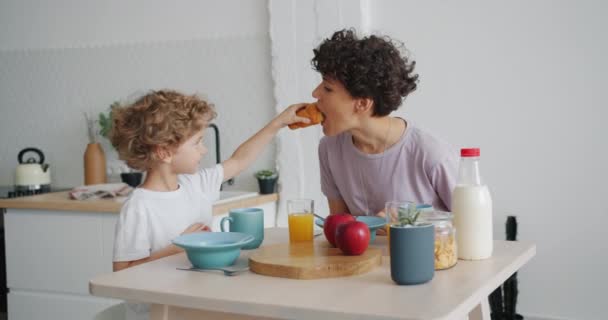 Niño pequeño alimentando a la madre con croissant y luego bebiendo durante el desayuno en casa — Vídeos de Stock