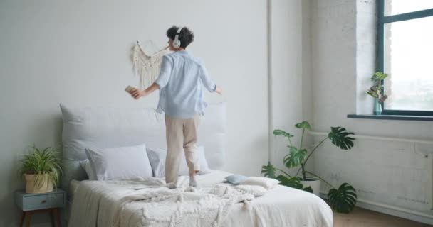 Весела дама насолоджується музикою в навушниках, тримаючи смартфон танцює на ліжку — стокове відео