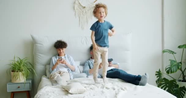 Vilain enfant sautant sur le lit tandis que les parents en utilisant des gadgets se détendre dans la chambre — Video