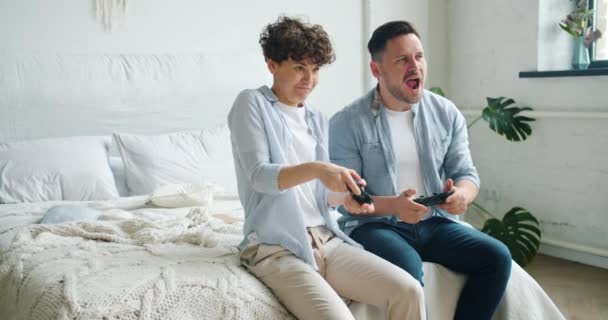 Chica y chico pareja disfrutando de video juego sentado en la cama en el apartamento de divertirse — Vídeos de Stock